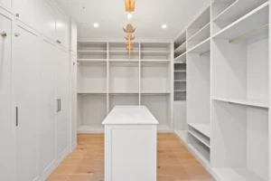 Ein geräumiger, moderner und maßangefertigter begehbarer Kleiderschrank mit weißen Regalen, Schränken und Hängestangen. Eine weiße Insel mit einer Marmorplatte steht auf einem hellen Holzboden. Die Deckenbeleuchtung umfasst eine dekorative Pendelleuchte aus Blattgold.