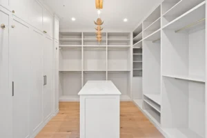 Ein geräumiger, moderner begehbarer Kleiderschrank mit weißen Einbauregalen und Schränken, eine weiße Mittelinsel, ein heller Holzfußboden und eine Deckenleuchte in Form eines Blattgoldes. Dieser Innenausbau erfolgt durch die Schreinerei Reinehr in Aachen