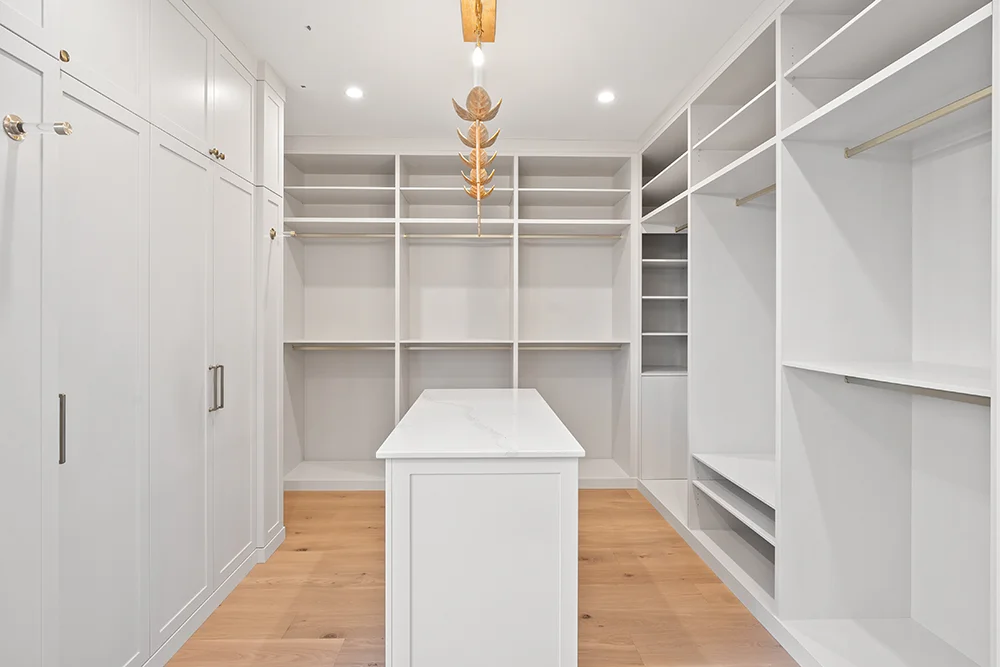 Ein geräumiger, moderner begehbarer Kleiderschrank mit weißen Einbauregalen und Schränken, eine weiße Mittelinsel, ein heller Holzfußboden und eine Deckenleuchte in Form eines Blattgoldes. Dieser Innenausbau erfolgt durch die Schreinerei Reinehr in Aachen