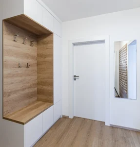 Moderner, minimalistischer Eingangsbereich mit weißen Wänden, einer weißen Tür, hellem Holzfußboden und einer eingebauten Bank mit Holzverkleidung, Schränken und Kleiderhaken. Ein Wandspiegel reflektiert einen Teil des Raumes.