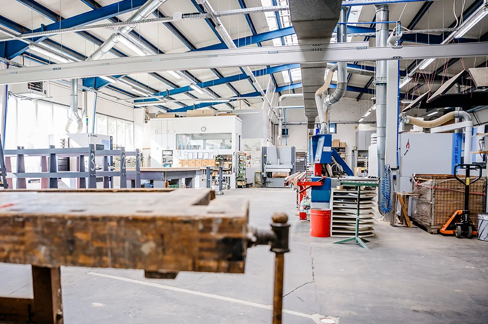 Eine geräumige, moderne Werkshalle der Schreinerei Reinehr in Aachen mit hohen Decken, industrieller Ausrüstung, Werkbänken, Regalen, Lüftungskanälen und großen Fenstern, die natürliches Licht hereinlassen. Der Bereich wirkt organisiert und sauber.