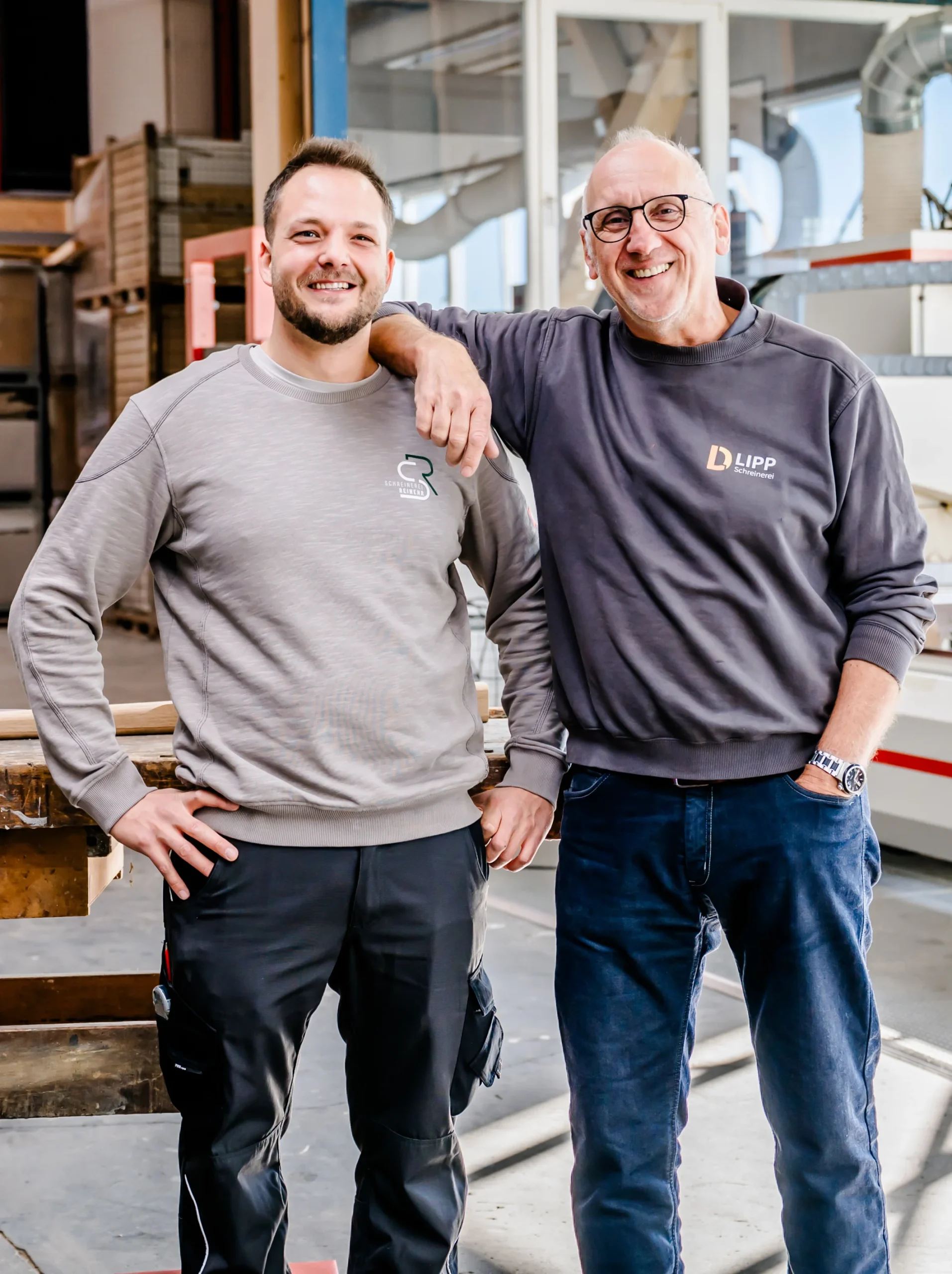 Felix Reinehr und ein Mann stehen lächelnd nebeneinander in einer hellen Industriehalle der Schreinerei, wobei der eine Mann seinen Arm auf die Schulter des anderen legt. Beide tragen legere Arbeitskleidung und Brillen.