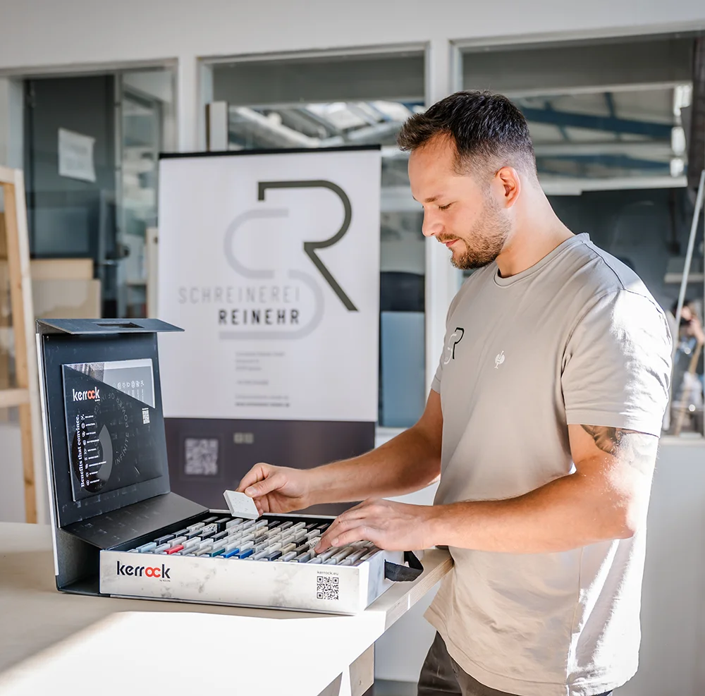 Handwerker Felix Reinehr steht in einer hellen Werkstatt und begutachtet eine mit "Kerrock" beschriftete Musterkiste mit verschiedenen Materialproben, während im Hintergrund eine Schautafel mit der Aufschrift "Schreinerei Reinehr" zu sehen ist.