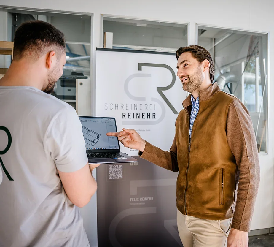 Felix Reinehr und ein Kunde stehen vor einem Laptop und sprechen über einen Entwurf für eine Maßanfertigung.