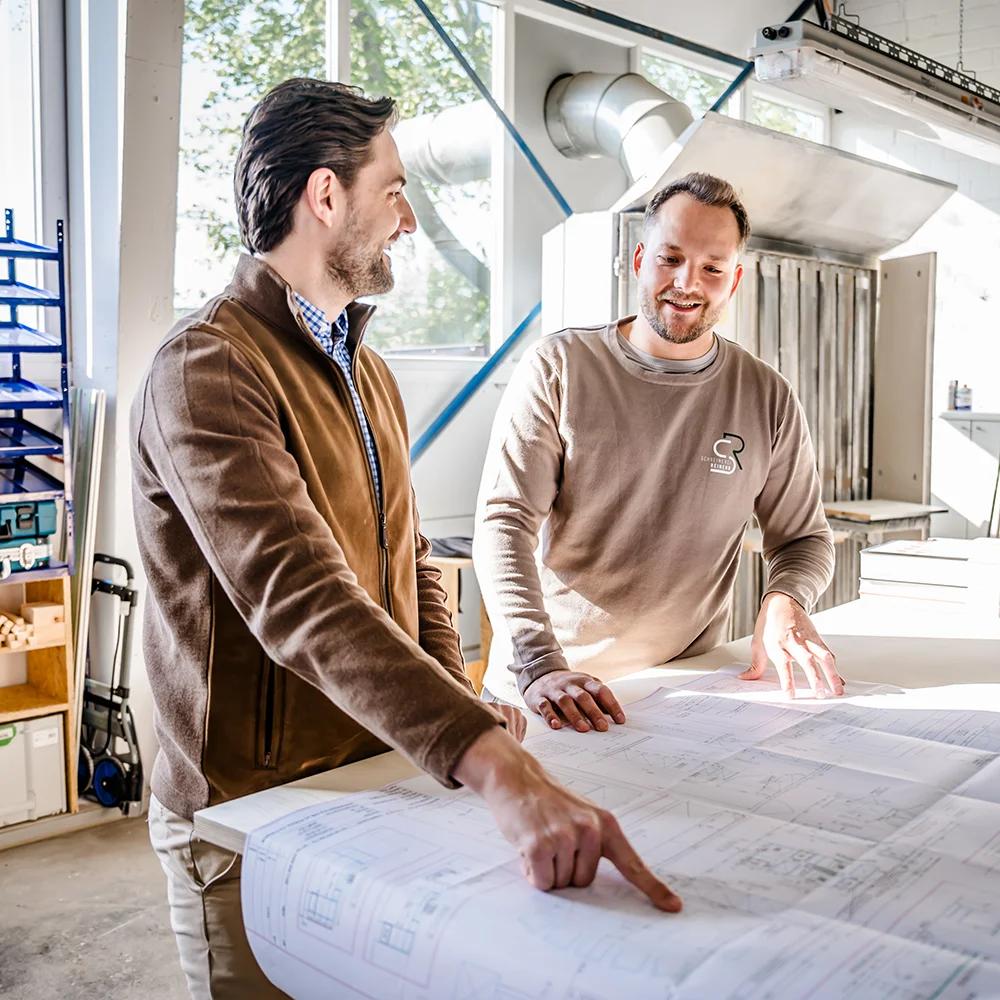 Felix Reinehr und ein Kunde stehen in einem hellen Büroraum und betrachten große Baupläne, die auf einem Tisch ausgebreitet sind, während sie miteinander diskutieren. Ein Mann zeigt auf einen Abschnitt auf den Plänen, während der andere zuhört und lächelt.